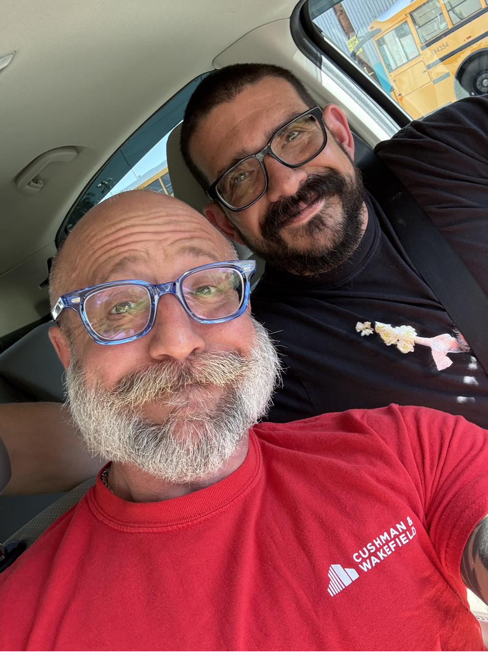 Barclay Barrios and a friend in a vehicle, both wearing glasses, smiling toward the camera.